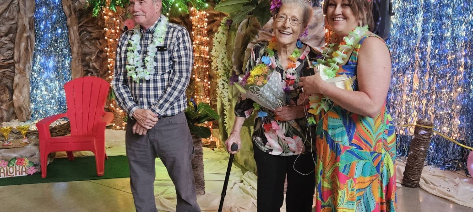 Three people at a tropical-themed party stand in front of a decorated backdrop with palm trees and string lights.